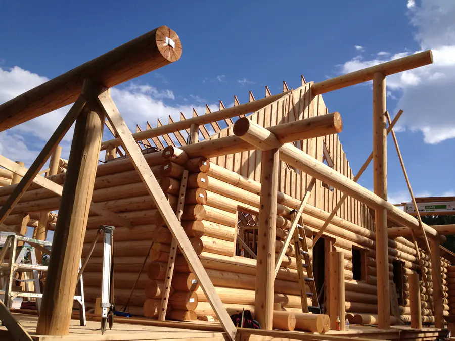 Round log walls going up: peeled logs stacked and numbered at the corner, and a log truss being set overhead against a blue sky.