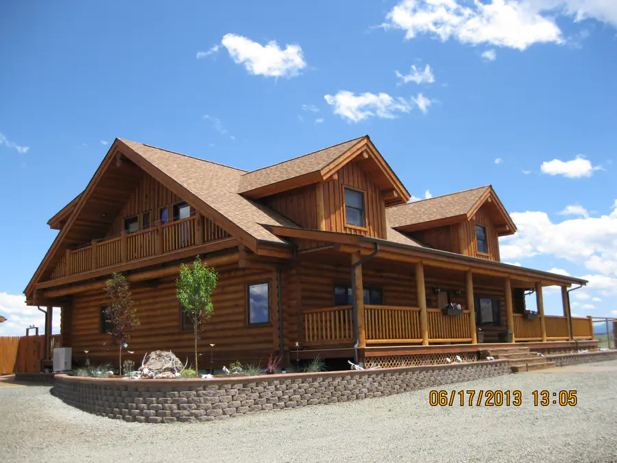 A round log home with dormers and a full-width porch, timbered gables and a stone retaining wall, under a blue sky.