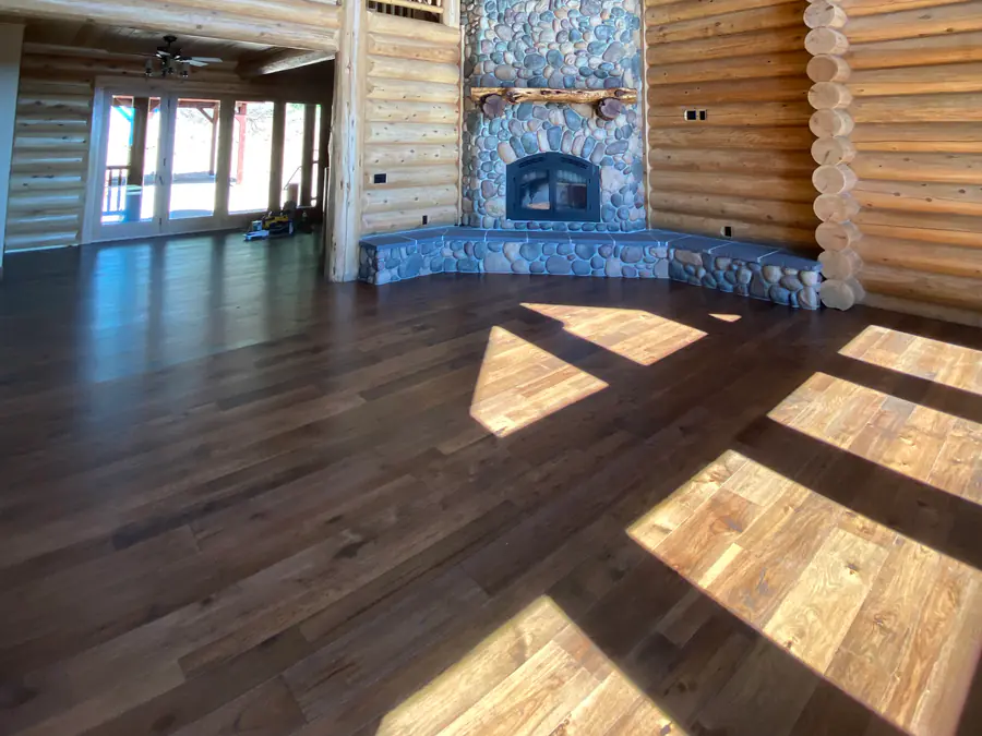 Inside a finished round log home: a stone fireplace, peeled log walls and tall windows, with sun across the wood floor.