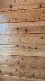Close on a hand hewn interior wall in warm honey tones: flat courses scalloped across their faces by the hewing tool, dark knots scattered along the grain, and a fine joint line between each course.