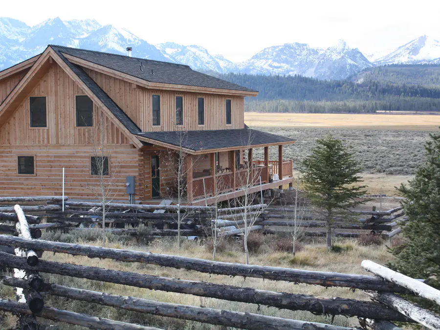 A two-storey squared timber home with a covered porch, on open ground before snow-capped mountains.