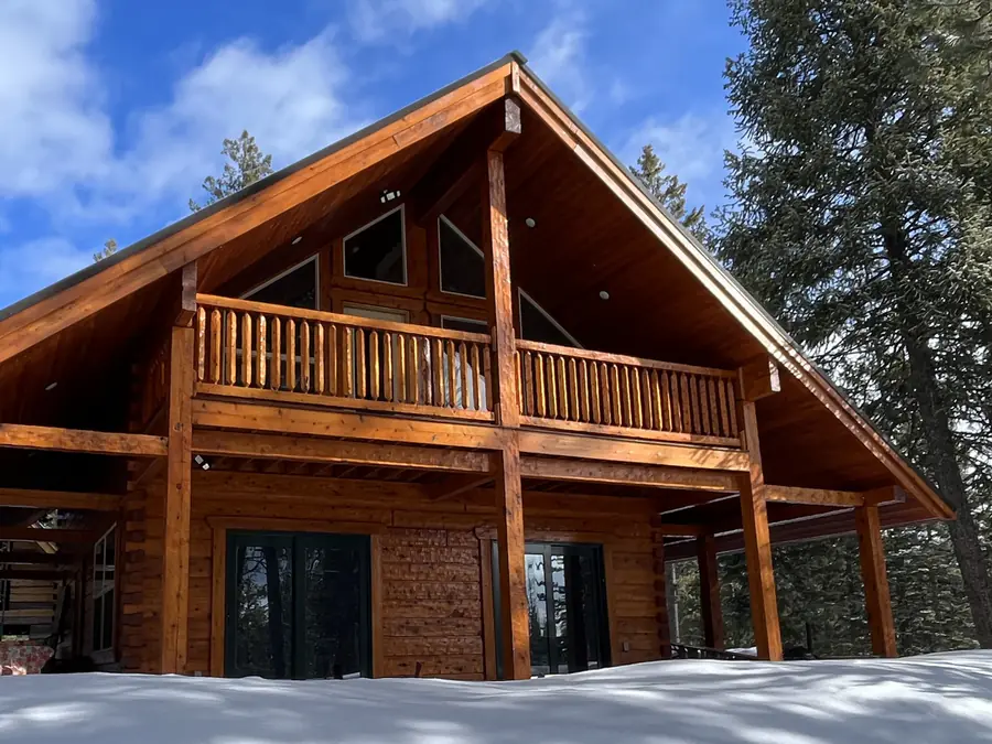A squared timber A-frame home with a wide deck on posts, in snow.