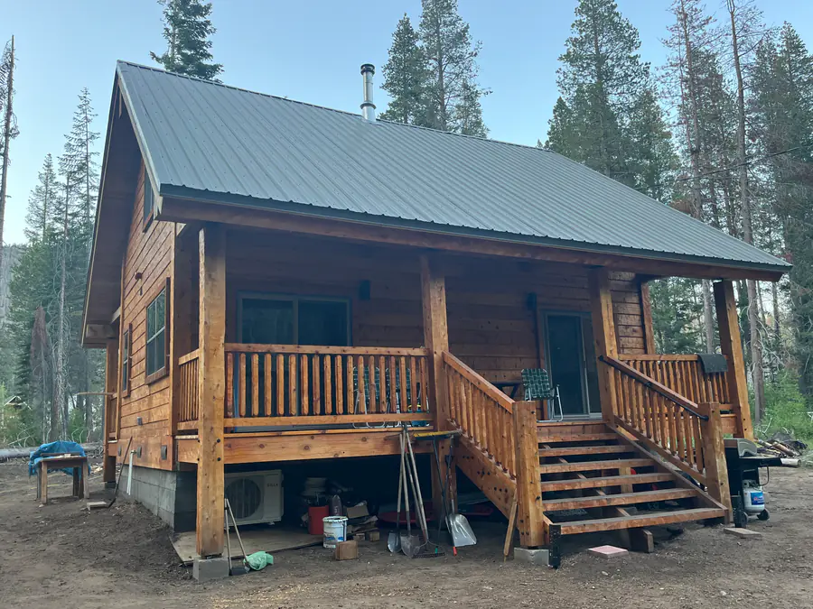 A small squared timber cabin with a green metal roof, a covered porch and log-post railings, among tall pines.