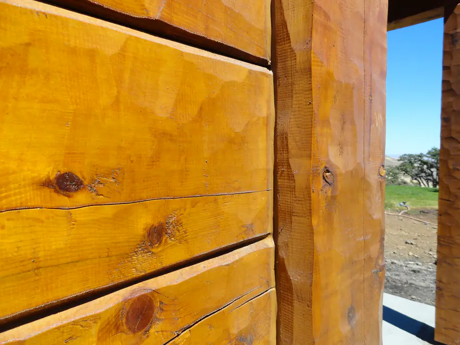 The hewn face of a squared cedar timber in close-up, its grain and a knot in full sun.