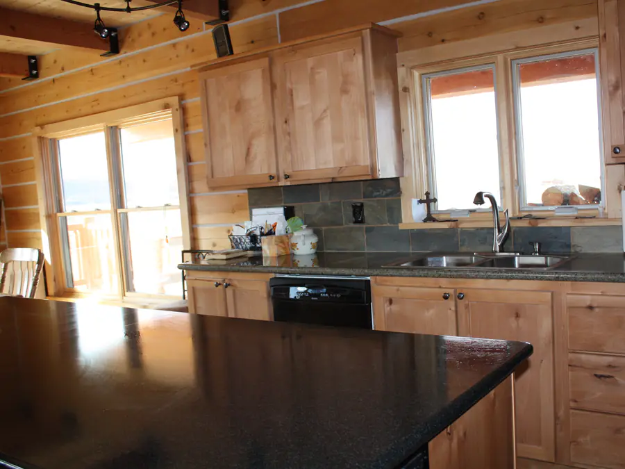 A kitchen inside a squared timber home, the hewn log wall behind the cabinets and a window over the counter.