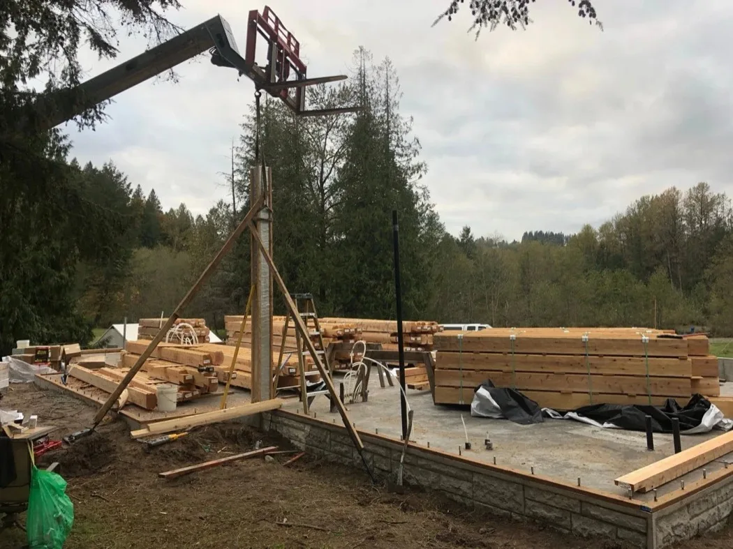A telehandler boom and a gin pole above banded cedar bundles stacked on a foundation slab, forest behind.