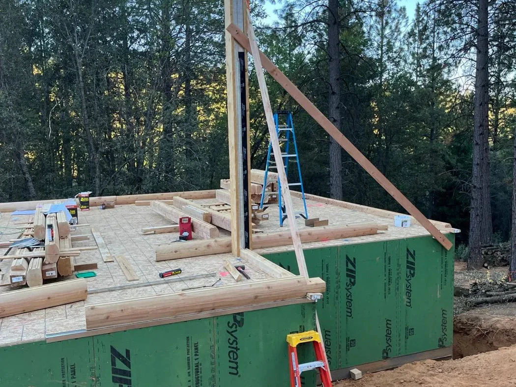 A subfloor over green sheathing with the first sill logs laid, a braced steel post standing at the opening and a ladder against it.
