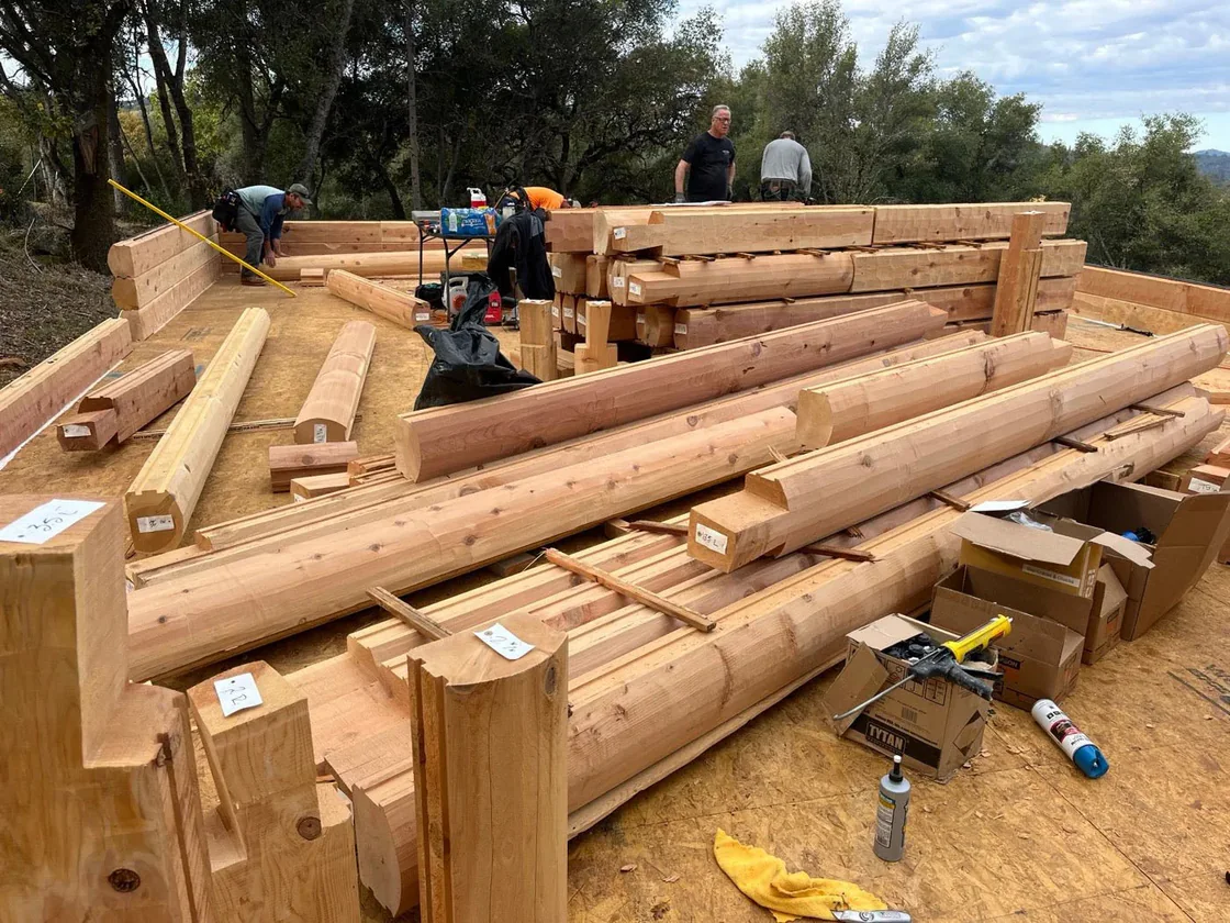 Sawn cedar logs and squared timbers laid out across a subfloor with their ends labeled, three men working among them.