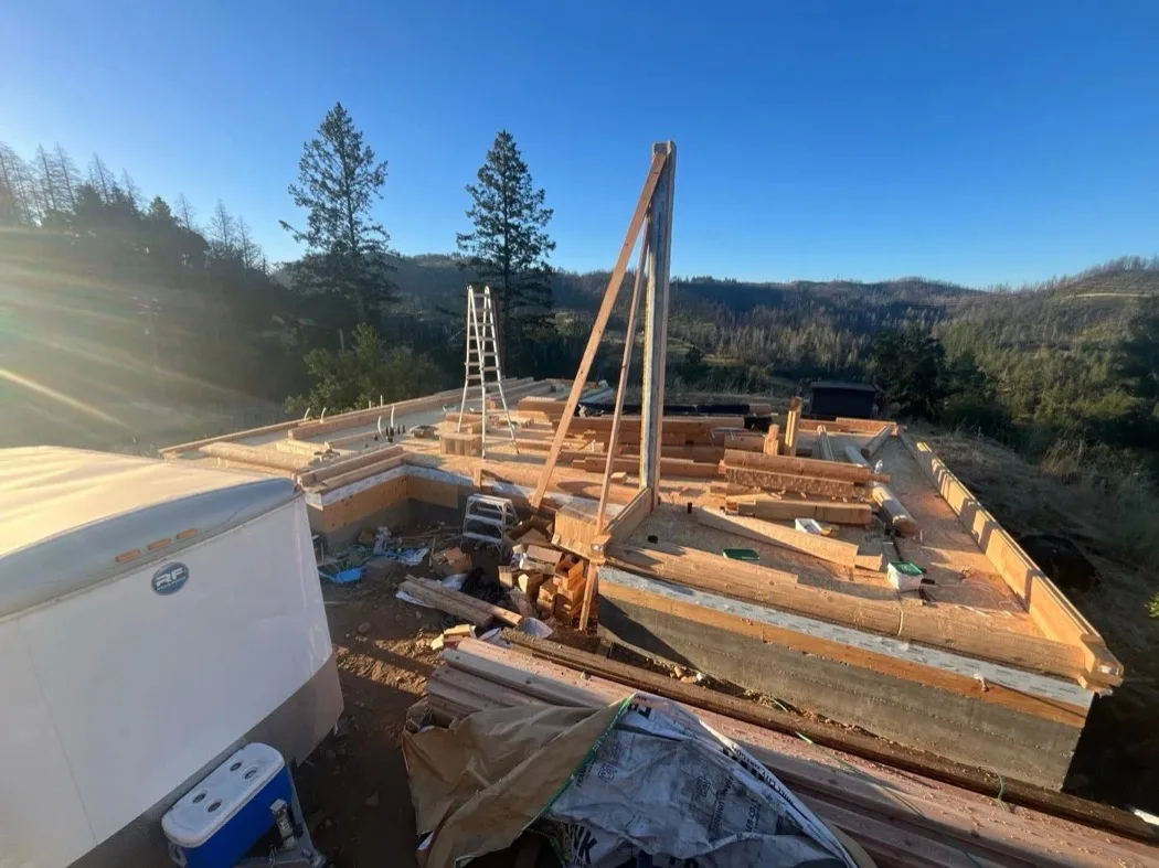 The first courses of a log wall on a foundation at sunrise, a site trailer and a cooler in the near corner and hills beyond.