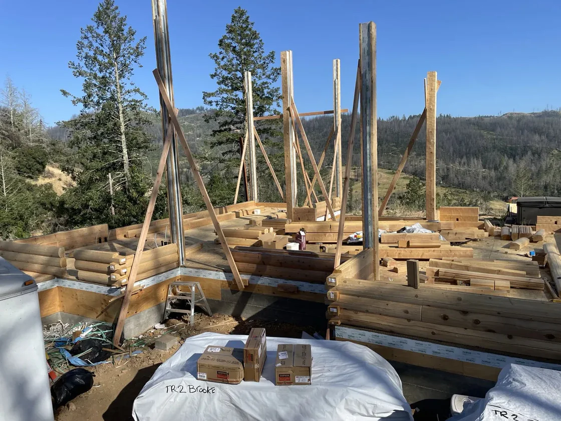 Log walls a few courses up with braced steel posts at the openings, wrapped bundles in the foreground and hills behind.