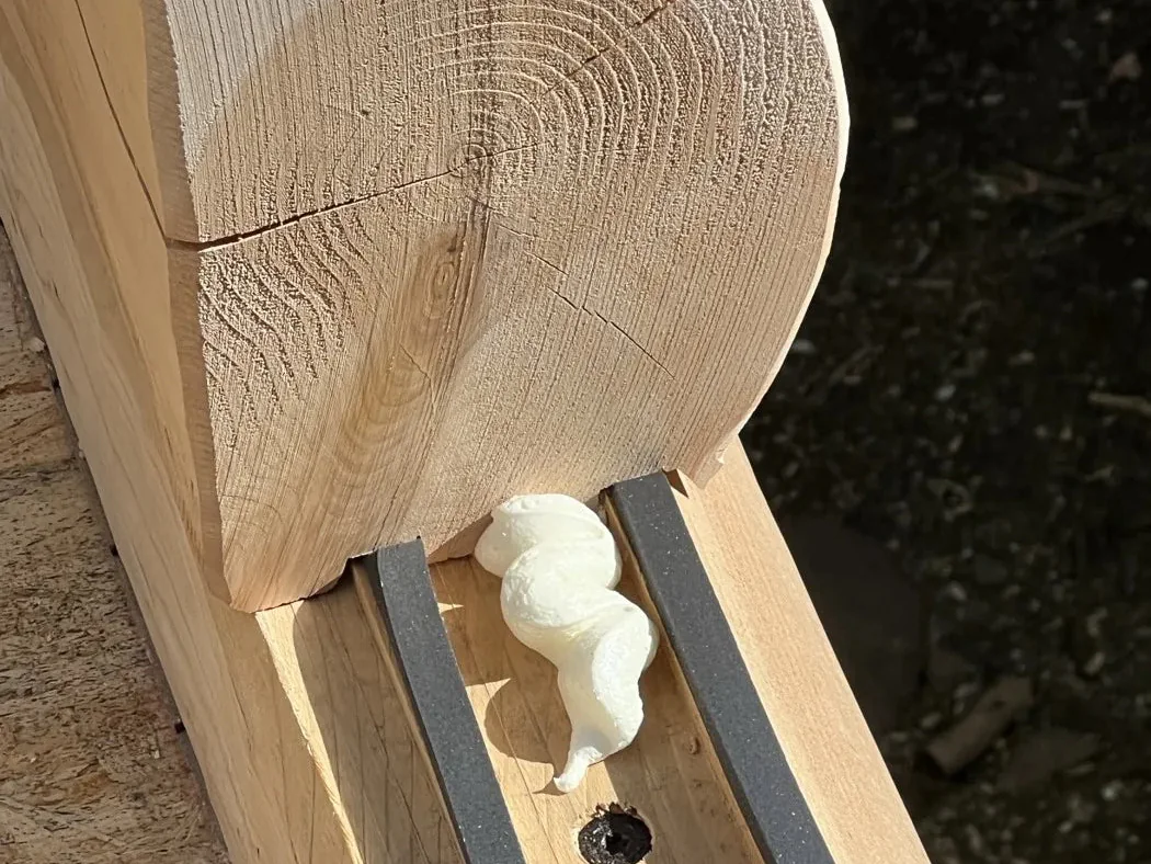The squared end of a cedar timber at a wall corner, its growth rings showing, with a bead of sealant laid along the joint.