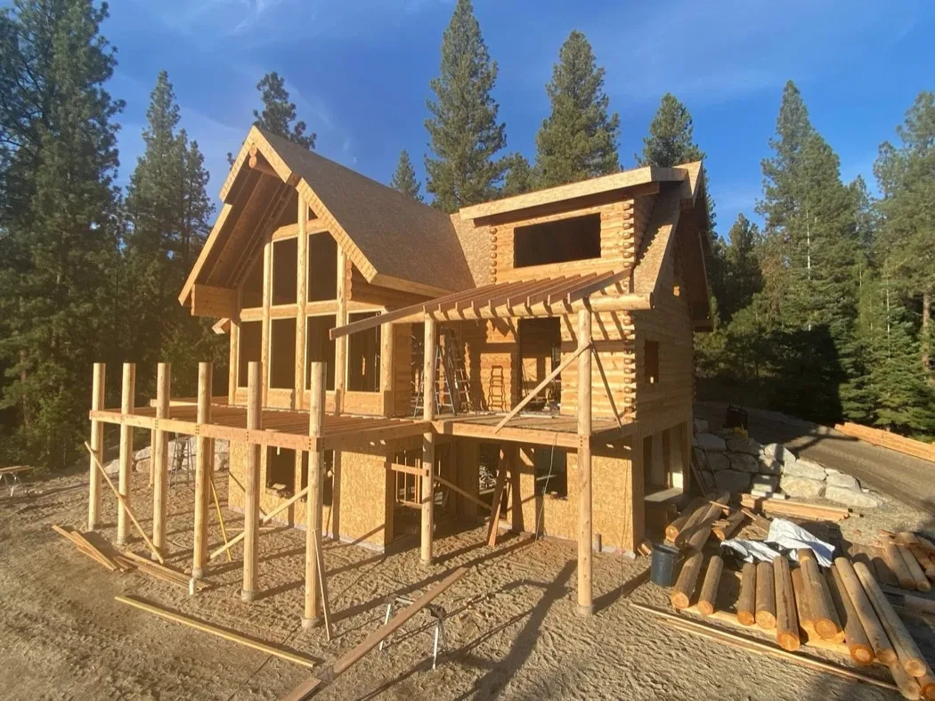 A two-story log shell at golden hour, deck posts up and sheathing still bare, with cut logs stacked on the ground.