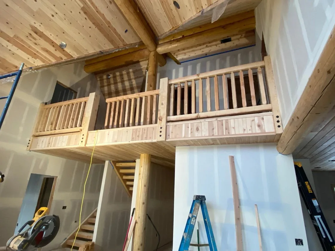 A loft landing inside an unfinished log home, log railings along its edge and the stair running down below.