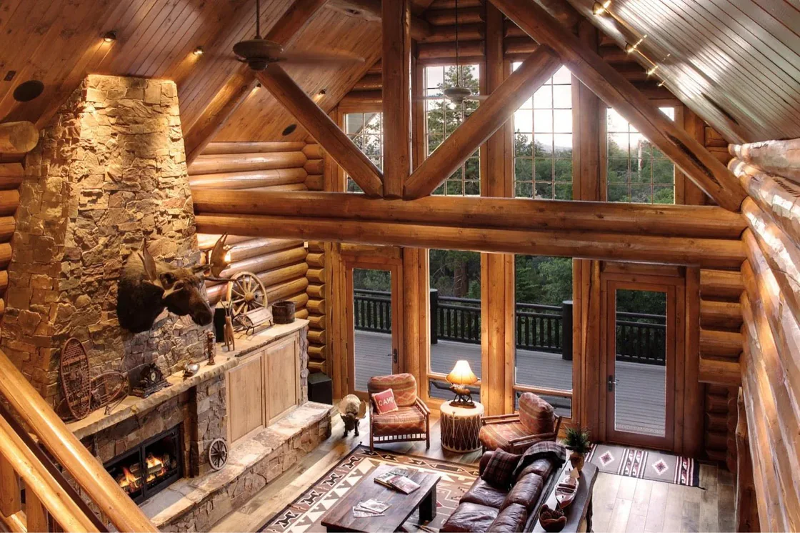 A great room in a finished log home: a stone fireplace rising to log trusses, leather sofas, and a wall of windows onto the trees.