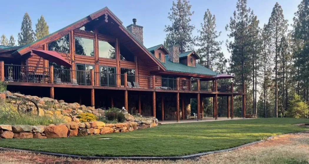 A finished two-story log home with a green metal roof and stone chimneys, a covered deck on log posts and a stone landscaping wall below a mown lawn.