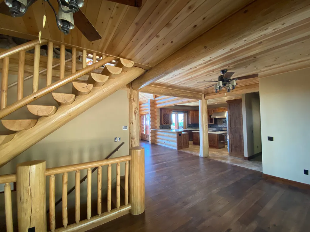 A log stair seen from the side in a finished log home: half log treads, their cut ends showing, climb along a peeled log horse under a cedar ceiling, with a log handrail and round log pickets above them. In front, a log railing and a log newel post guard the stair down to the floor below.