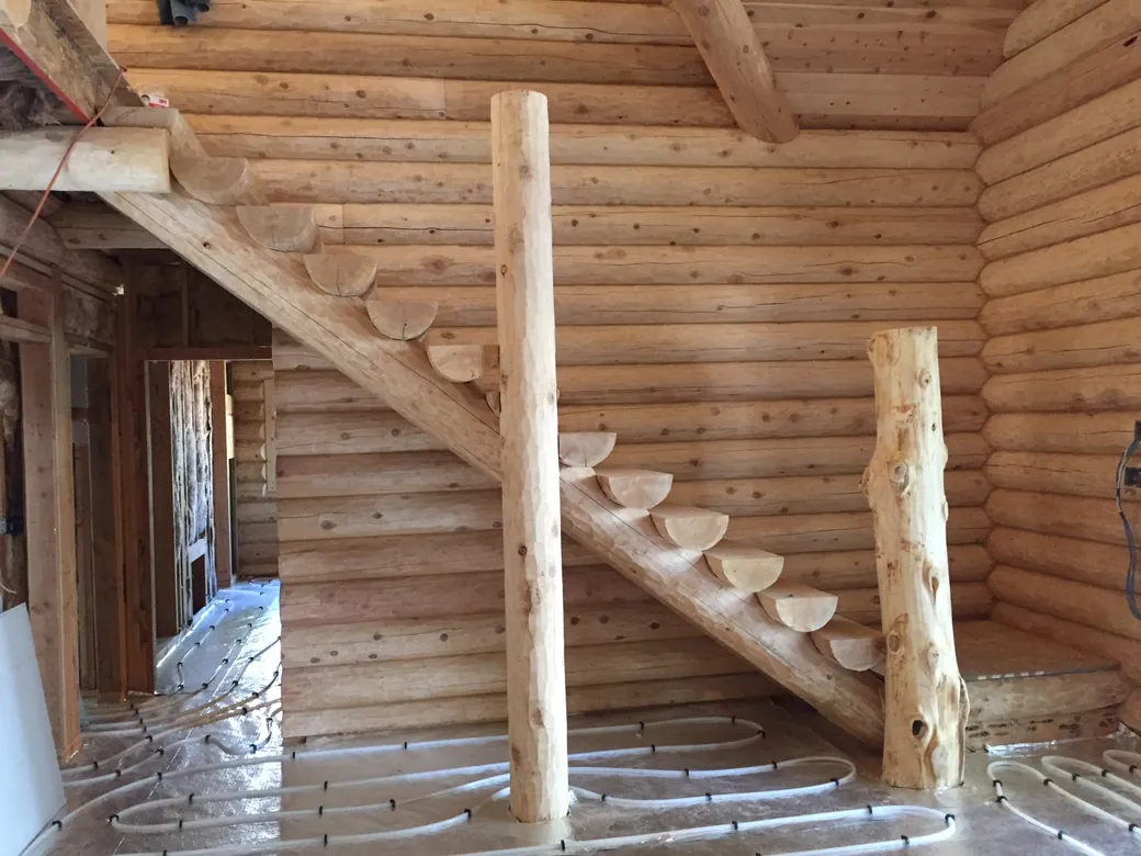 A log stair climbing a round log wall in a house still being built: half log treads, cut ends showing, along a peeled log horse, with a peeled log post and a knotty natural log post standing in front and radiant heat tubing looped across the floor.