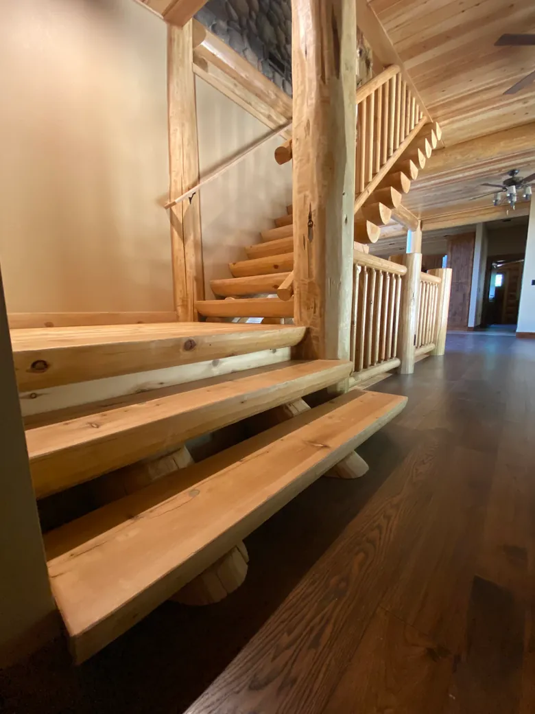 Low at the foot of a log stair: three half log treads, flat side up and sanded smooth, rest across round log horses whose ends show beneath them. Beyond, the stair turns and climbs past a peeled log post, its half log treads in a row under a log railing.