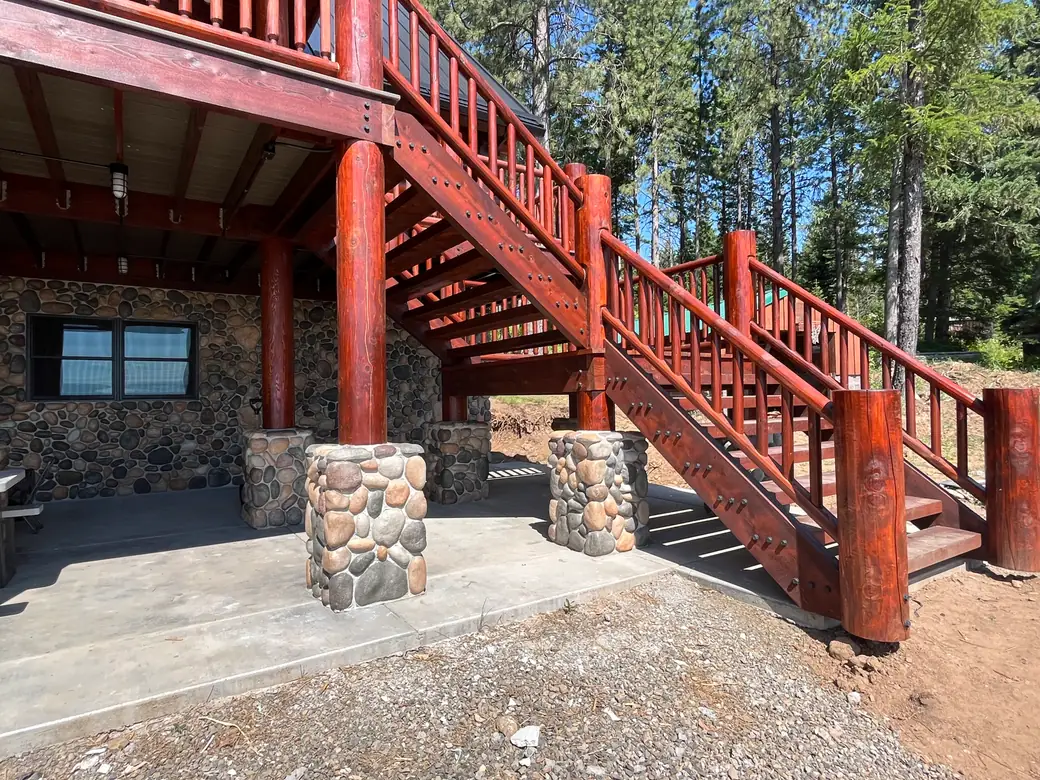 A two-flight outside stair up to a covered deck, stained deep red: timber horses with the treads between them and rows of black hex nut heads down their faces, log posts at the landing and the foot, and log railings with round pickets up both sides. River rock piers and a concrete patio under the deck, pines behind.