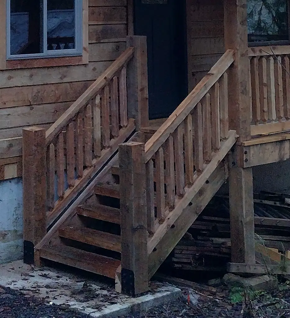 A short outside stair up to a front door at dusk: timber horses and treads, square timber posts at the foot, black at their base, timber handrails over shaped timber pickets, and the same pickets along the porch railing beside it.