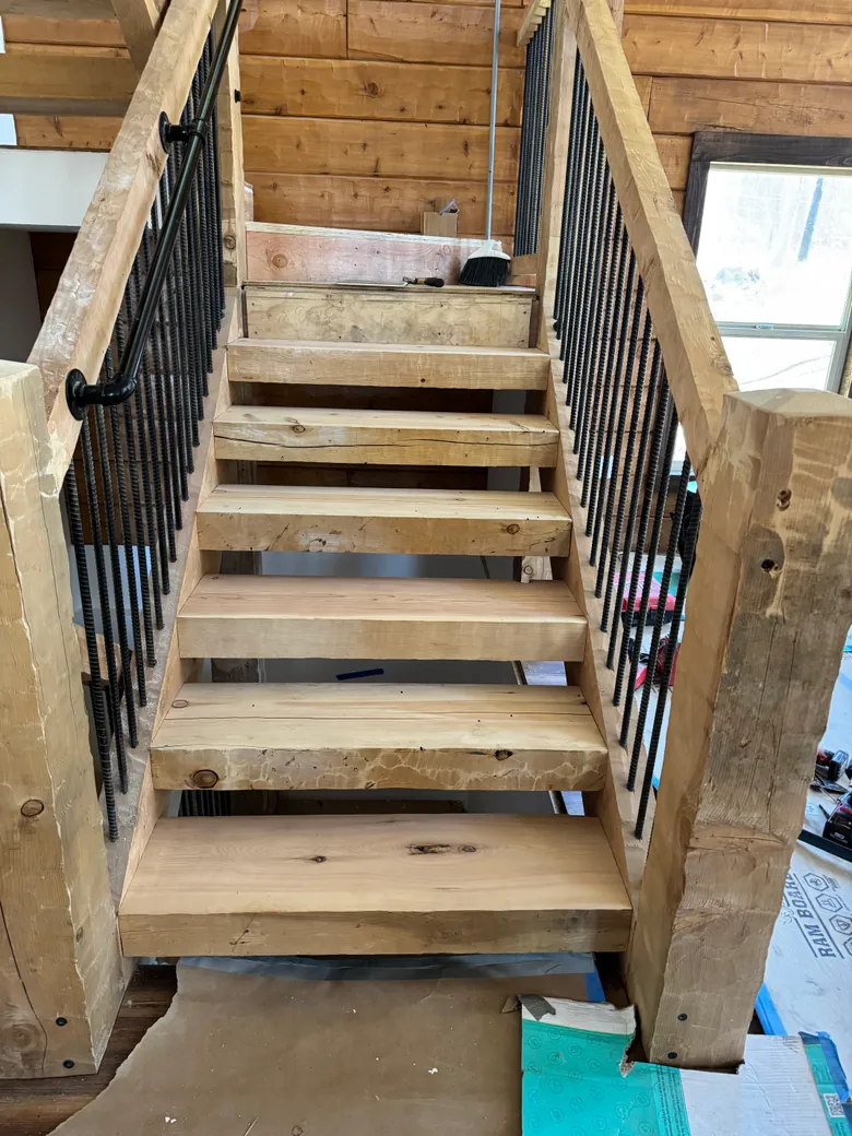 Looking up an indoor stair of thick, hand hewn timber treads set between timber horses, square hewn newel posts at the foot, timber handrails over black rebar pickets and a black pipe handrail on the left, with paper still laid over the floor.